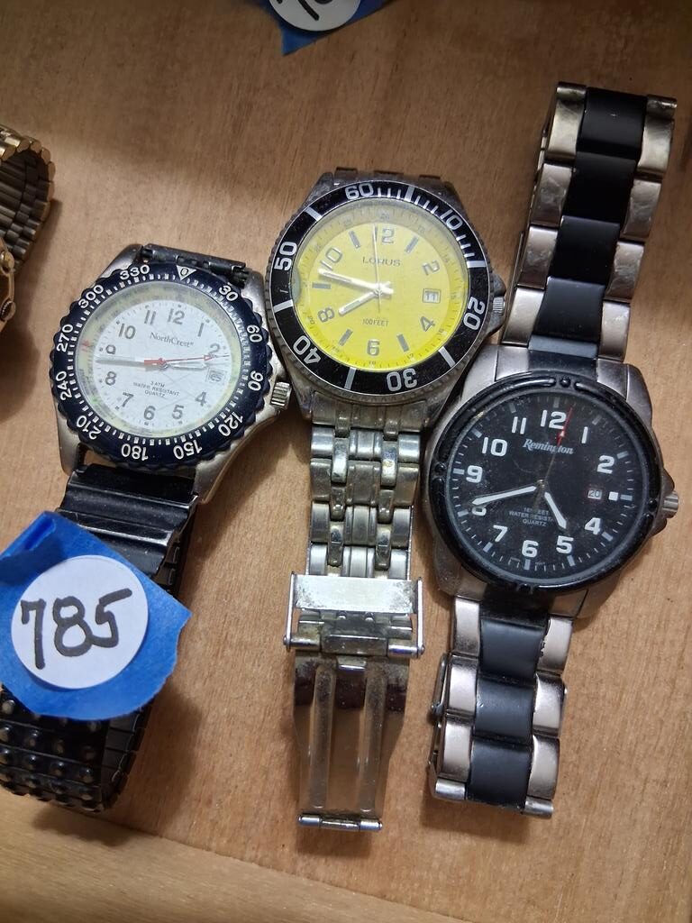 Auction Item Three men’s wristwatches with metal bands displayed on a wooden surface, including black, yellow, and white-faced designs. Las Vegas Auctions