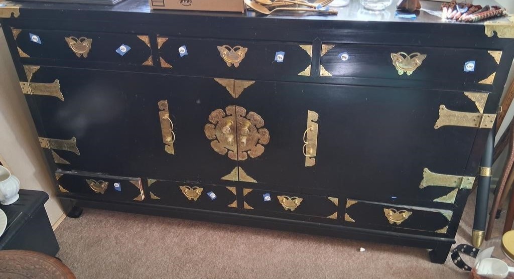 Auction Item Black lacquered Asian chest with ornate brass hardware, featuring decorative hinges, butterfly-shaped corner plates, and a circular center medallion clasp. Las Vegas Auctions